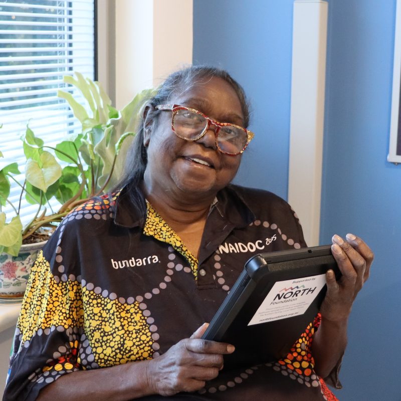 A woman in front of a window is smiling as she looks at the iPad she is holding.