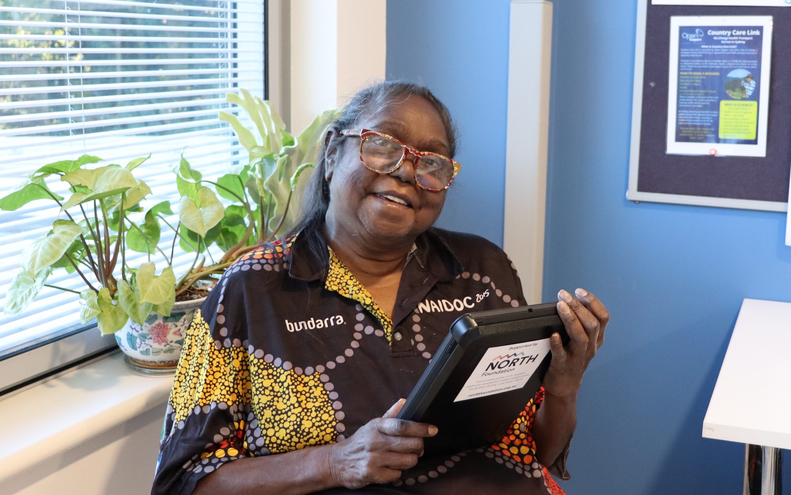 A woman in front of a window is smiling as she looks at the iPad she is holding.