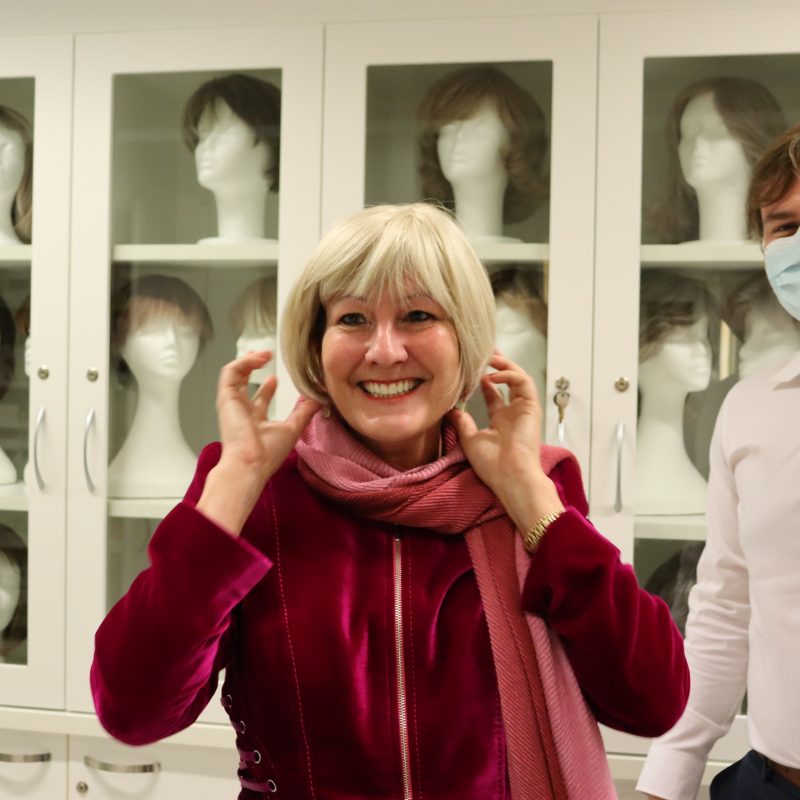 Woman trying short blond wig, man wearing mask in background