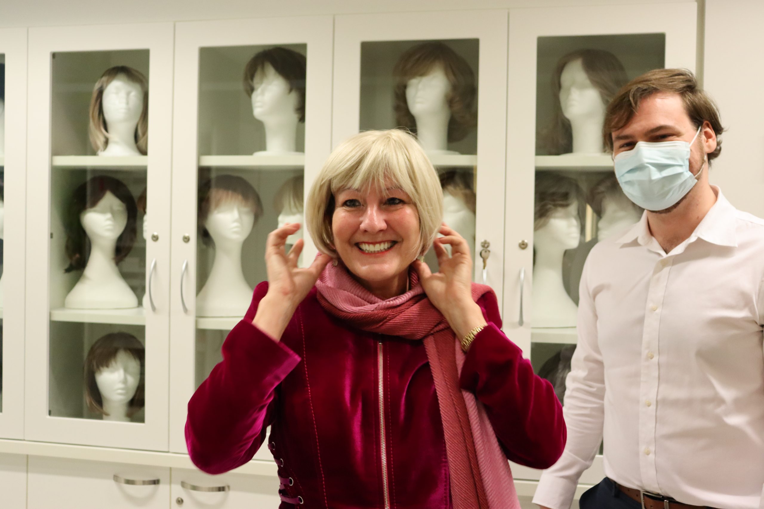 Woman trying short blond wig, man wearing mask in background